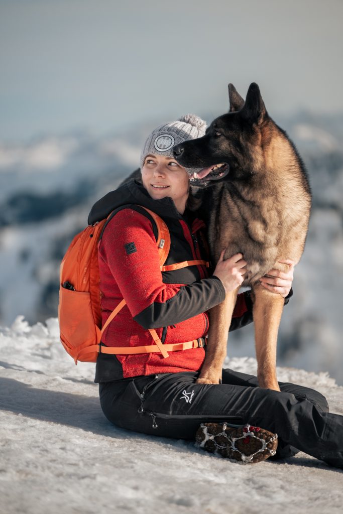 Person sitzt mit Hund im Schnee am Hochgrat im Allgäu und hält ihn beim Wandern mit Hund in den winterlichen Bayerischen Alpen